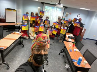 Mask wearing mult-cultural group of women and one man holding similar paintings with a vitrual group in the background located in a conference room and the instructor, a woman with spectacles, brown skin and hair wrap in the foreground