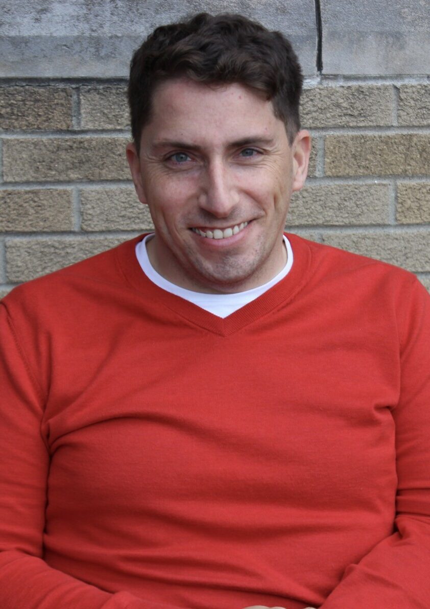 A headshot of Paul smiling at the camera. he is wearing a red shirt.
